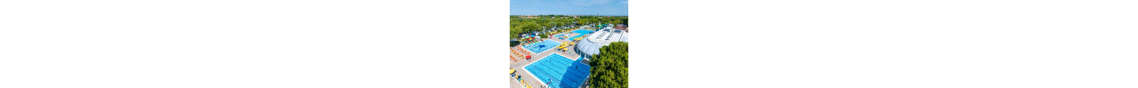 A large outdoor swimming pool complex is captured from above on a sunny day. Multiple blue pools, including a main lap pool and a shallow children’s pool with play structures, are surrounded by sun loungers and colorful umbrellas. A white domed building and several water slides add to the recreational atmosphere. Lush green trees and a nearby campground or holiday park frame the aquatic area, suggesting a busy family vacation destination.