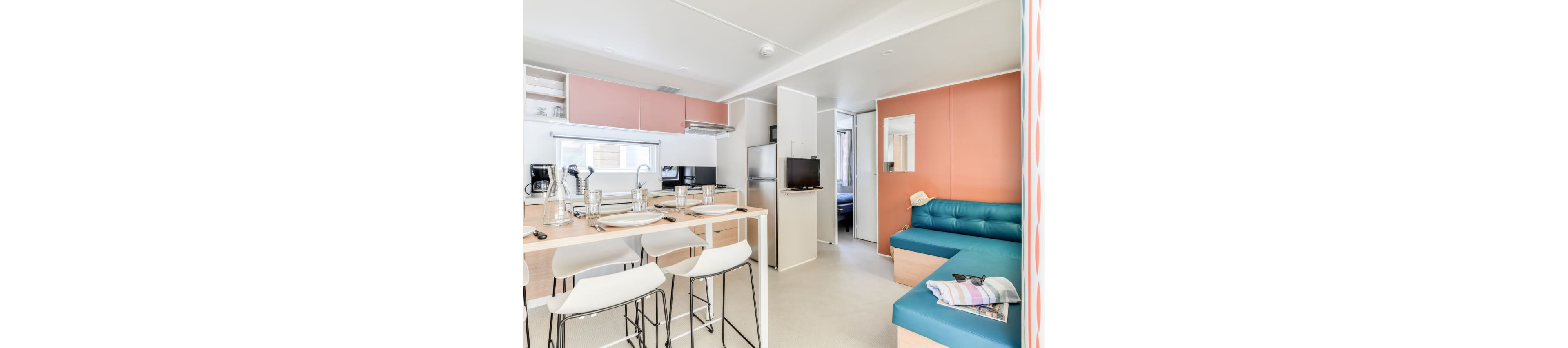 A compact modern studio features an open-plan kitchen and living space. The kitchen includes a wooden island with bar stools, white dishes, and stainless steel appliances. A teal sofa sits against a warm coral accent wall, with folded towels placed on the arm. Large windows and light-colored surfaces create an airy, minimalist atmosphere, while a small TV and visible bedroom doorway suggest a vacation rental or small apartment layout.