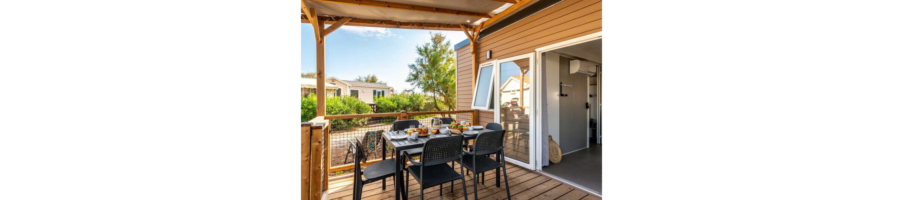 A wooden terrace features a dining table set for a meal outside a modern cabin or mobile home. Black chairs surround the table, which is arranged with plates, glasses, and assorted breakfast foods. The covered patio overlooks greenery and neighboring cabins under a bright blue sky. Sliding glass doors open into a simple, contemporary interior, suggesting a relaxed vacation setting.