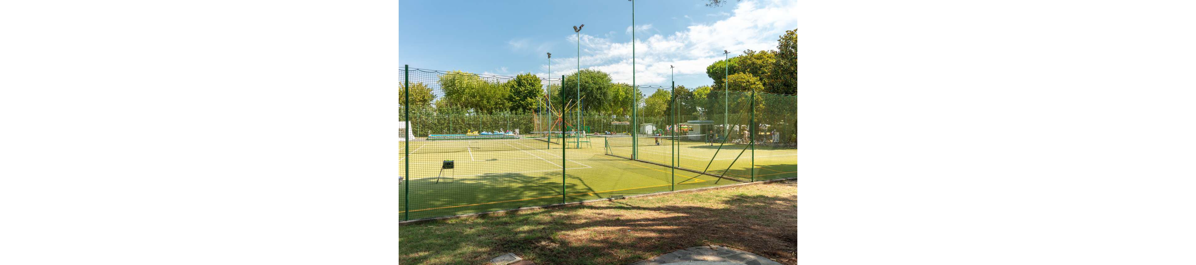 An outdoor tennis court is enclosed by a tall green fence in a sunny park setting. The court surface appears well-maintained with clear white lines and a central net. Trees and greenery surround the sports area, with additional recreational structures visible in the background. Bright daylight and blue sky create a warm, inviting atmosphere for outdoor activities.