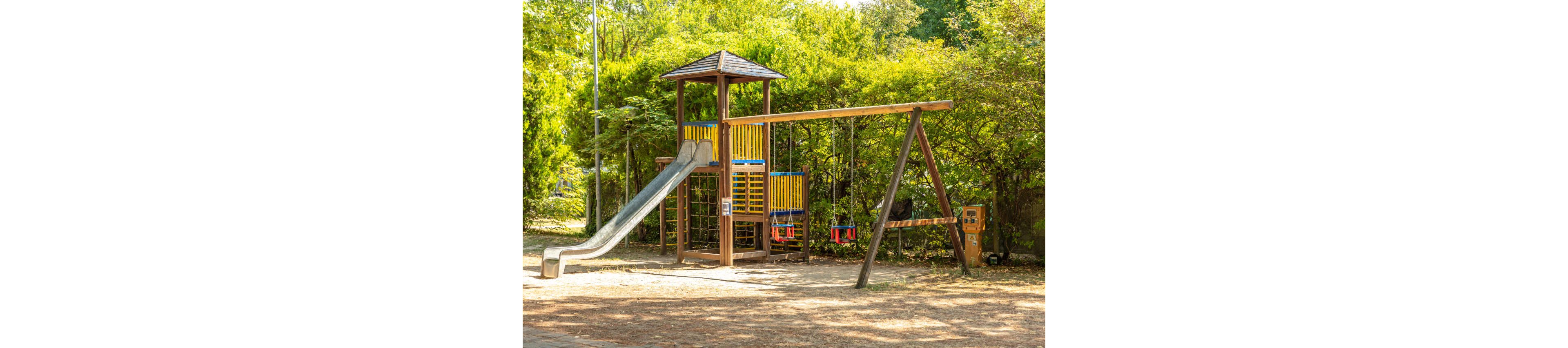 A wooden playground structure stands in an outdoor park, featuring a metal slide, climbing frame, and colorful railings. Two empty swings hang from a wooden swing set beside the main tower. The ground is covered with dry dirt and scattered leaves, suggesting a warm, sunny day. Lush green trees and bushes surround the play area, creating a natural, shaded backdrop.
