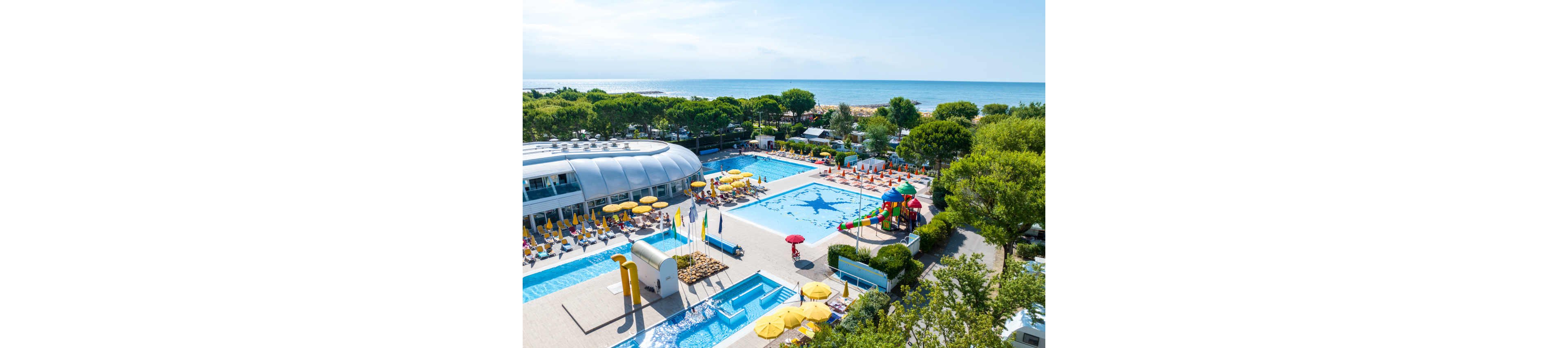 A large family-friendly resort features multiple outdoor swimming pools near a sandy beachfront. Colorful sun loungers and yellow umbrellas surround the main pool, while a children’s splash area with slides and play structures adds fun for younger guests. Modern low-rise buildings and lush green trees frame the aquatic area, creating a relaxed holiday atmosphere. In the distance, the calm sea and clear sky complete the coastal vacation setting.