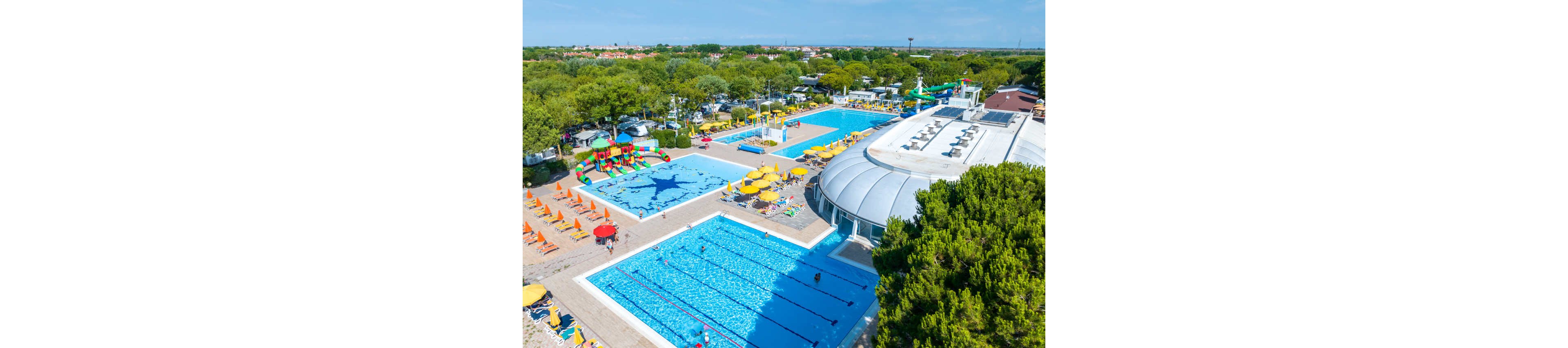 A large outdoor swimming pool complex is captured from above on a sunny day. Multiple pools, including a lap pool and a shallow leisure pool with a star design, are surrounded by sun loungers and umbrellas. A domed building and water play structures add interest to the recreational setting. Lush green trees and a distant townscape frame the vibrant holiday atmosphere.