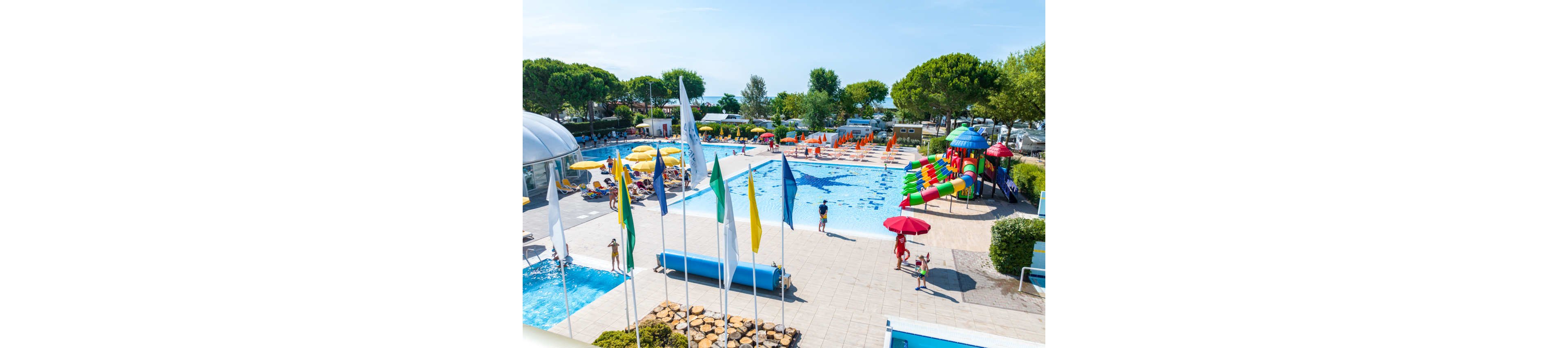 Large outdoor swimming pools are surrounded by sun loungers and colorful umbrellas at a busy holiday resort. Children and adults enjoy the water, slides, and splash features under a bright blue sky. Tall trees and landscaped areas frame the pool deck, while multiple flags wave in the foreground. The scene conveys a lively, family-friendly vacation atmosphere with vibrant colors and clear weather.