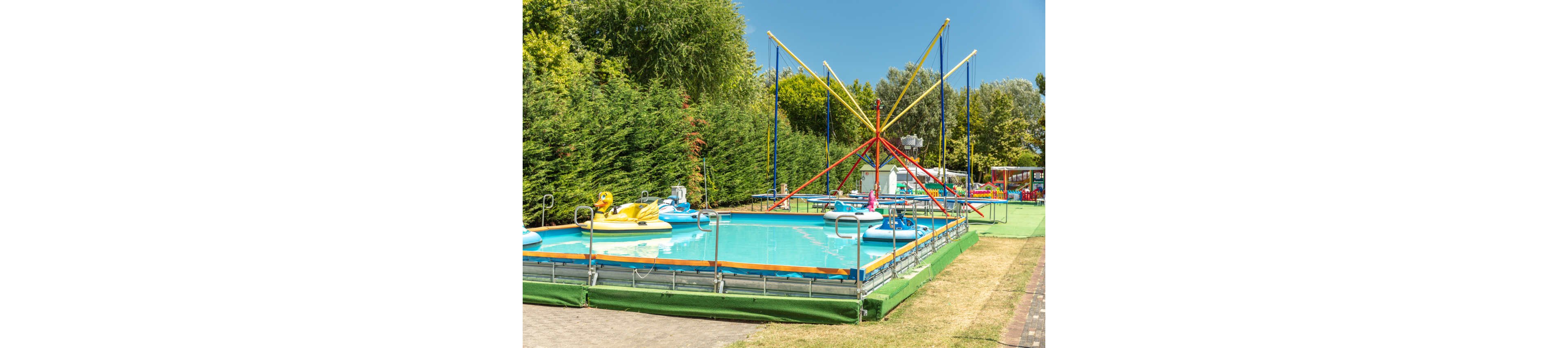 An outdoor amusement area features a shallow rectangular pool with colorful paddle boats floating on the water. A bungee trampoline structure with tall poles and elastic cords stands beside the pool. The scene is surrounded by dense green hedges and trees under a clear blue sky. Bright colors and sunny weather create a cheerful, family-friendly atmosphere.