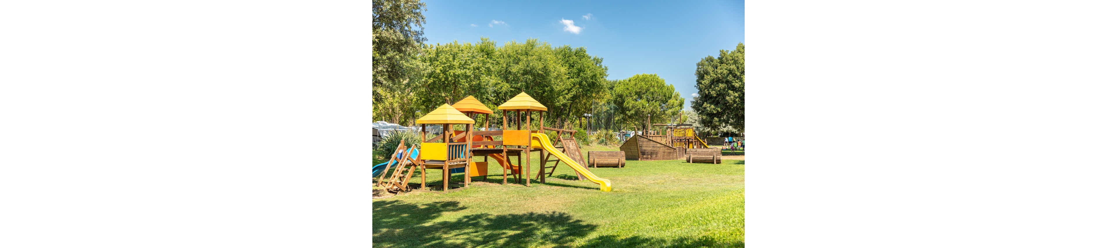 A colorful outdoor playground with wooden climbing frames and yellow slides stands on a large grassy field. Multiple play structures, including towers and a ship-like climbing area, are spread across the park. Lush green trees surround the playground, providing a natural backdrop under a bright blue sky. The scene suggests a sunny, family-friendly recreational area for children.