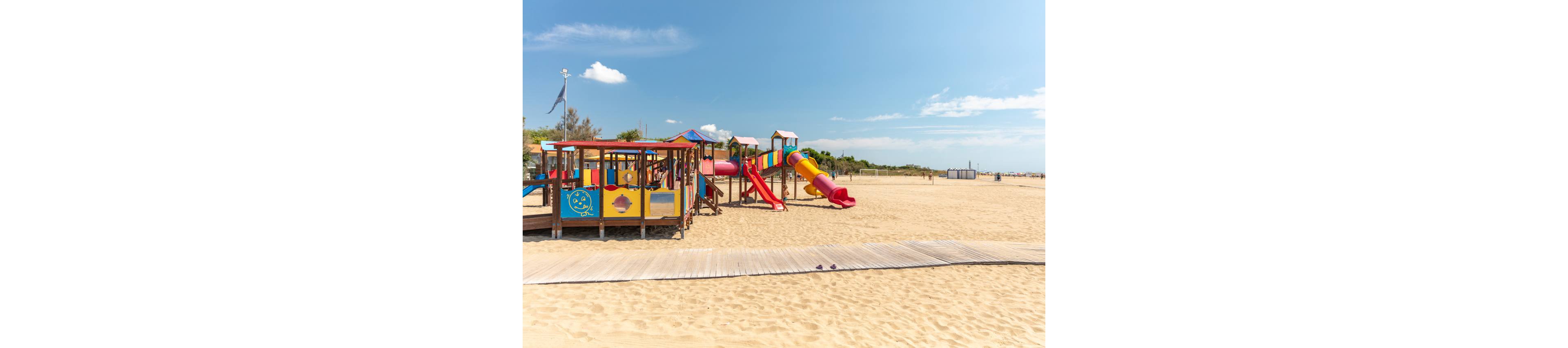 A brightly colored playground stands on a wide sandy beach under a clear blue sky. The play area features slides, tunnels, and climbing structures in red, yellow, blue, and green. Wooden walkways cross the sand in the foreground, leading toward the play equipment. In the distance, low vegetation and beach facilities line the horizon, emphasizing the open, relaxed seaside setting.