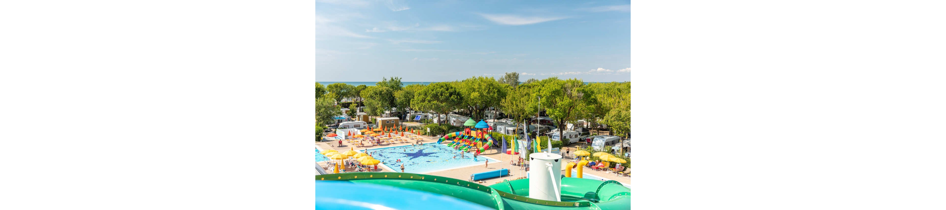 A large outdoor swimming pool complex is surrounded by colorful slides and play structures in a family-friendly campsite. People relax on sun loungers under yellow umbrellas while others swim and play in the pool. Caravans, tents, and camper vans are visible among the trees around the water park. In the distance, the blue sea and clear sky create a bright summer holiday atmosphere.