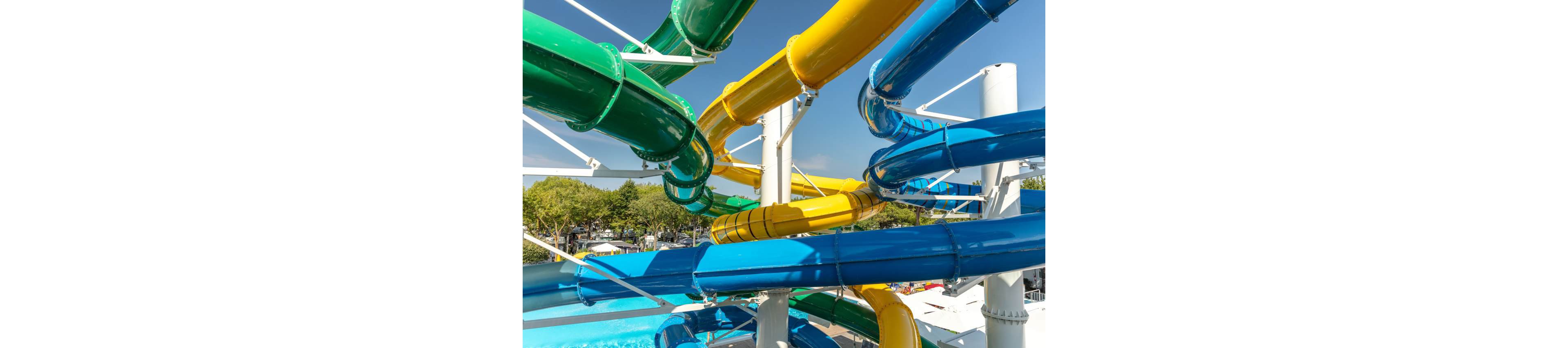 Brightly colored water slides twist and overlap in an outdoor water park. Large green, yellow, and blue tubes curve around a central white support structure. Sunlight highlights the glossy surfaces of the slides against a clear blue sky. Trees and park buildings are visible in the background, suggesting a busy recreational area.