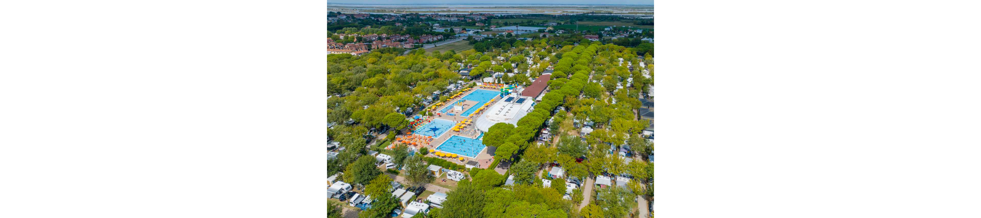 A large campsite resort with multiple swimming pools is nestled within a dense forest of green trees. The aerial view shows bright blue pools surrounded by yellow sun loungers and umbrellas, with white camper vans and tents arranged in neat rows around the complex. A central pool features a star-shaped design and a shallow play area, suggesting family-friendly recreation. In the distance, a lagoon or coastal waterway and a small town are visible, adding to the holiday atmosphere.
