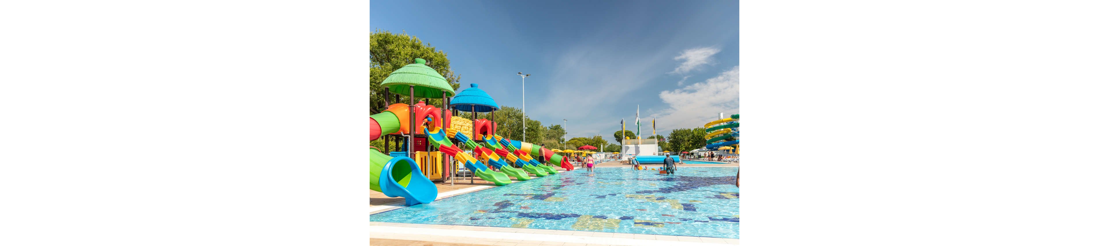 A large outdoor water park features multiple colorful slides emptying into a shallow pool. Children and adults wade and play in the clear blue water under a bright sunny sky. The play structures include green-roofed towers, red and yellow slides, and a curved blue tube slide. Trees, flags, and additional slides are visible in the background, creating a lively summer recreation scene.
