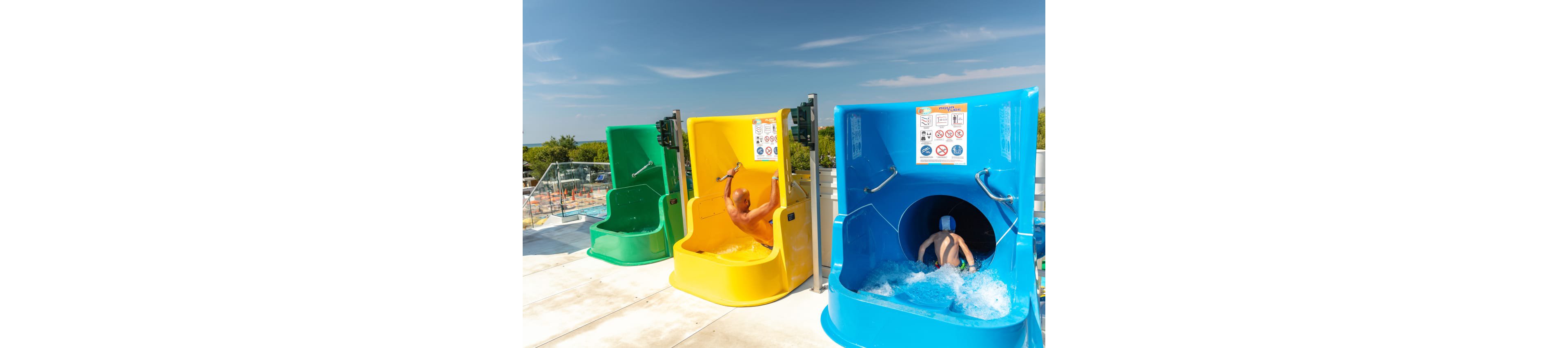 Two children sit in bright yellow and blue launch pods at the top of a water slide attraction. One child prepares to drop while the other splashes into the enclosed blue slide. A third empty green slide pod is visible beside them. Safety instruction signs are posted above each slide, and a sunny outdoor water park with trees and pools appears in the background.