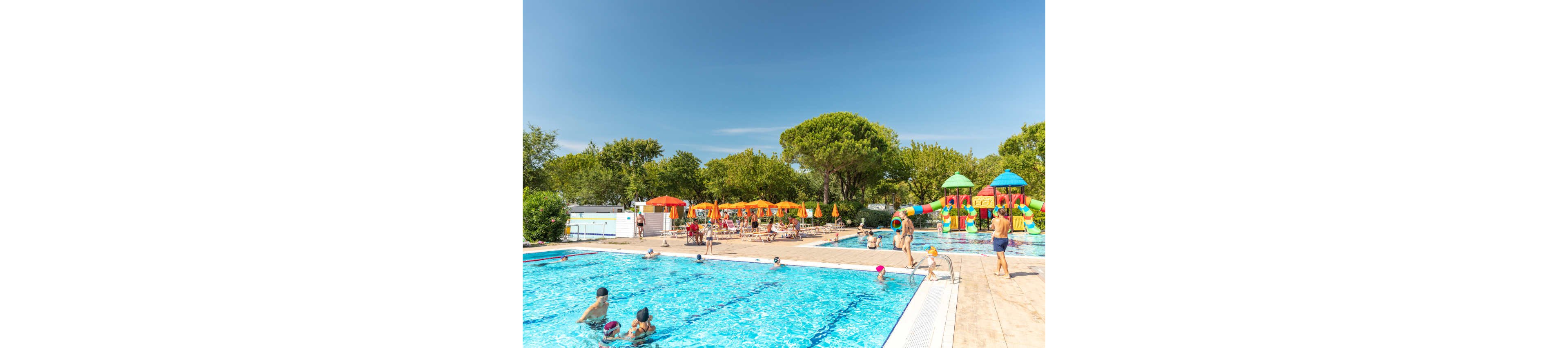 An outdoor swimming pool is filled with people enjoying a sunny day. Families and children play in the clear blue water while others relax on the poolside deck. In the background, colorful umbrellas and lounge chairs provide shaded seating areas. A bright water playground with slides and towers adds a fun attraction for kids, all set among green trees under a clear blue sky.