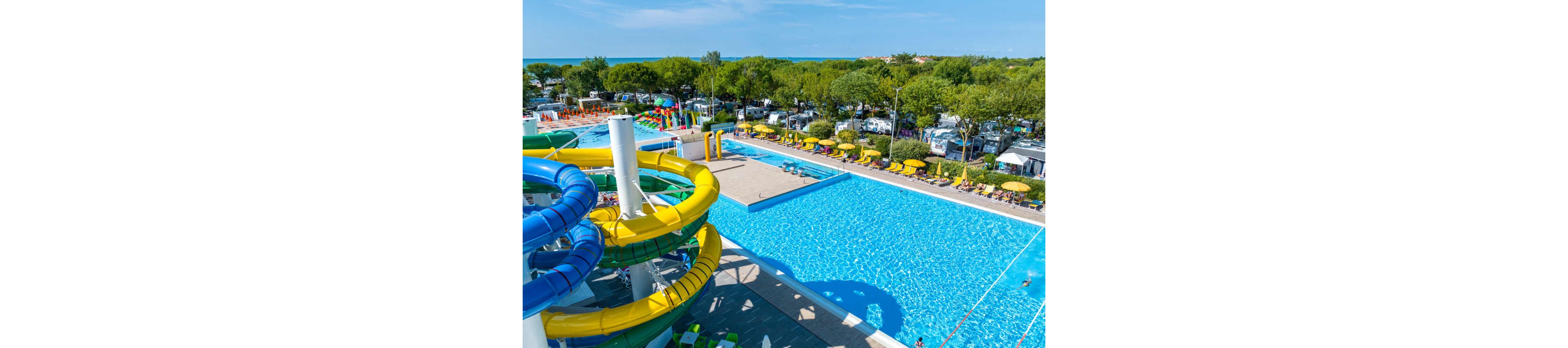 A spacious outdoor swimming pool complex features bright yellow and green water slides in the foreground. Sun loungers and umbrellas line the poolside, with people relaxing and enjoying the sunny day. Beyond the pool area, a campground or holiday park with trees and parked vehicles stretches toward the horizon. In the distance, the blue sea is visible under a clear sky, creating a vibrant summer vacation atmosphere.