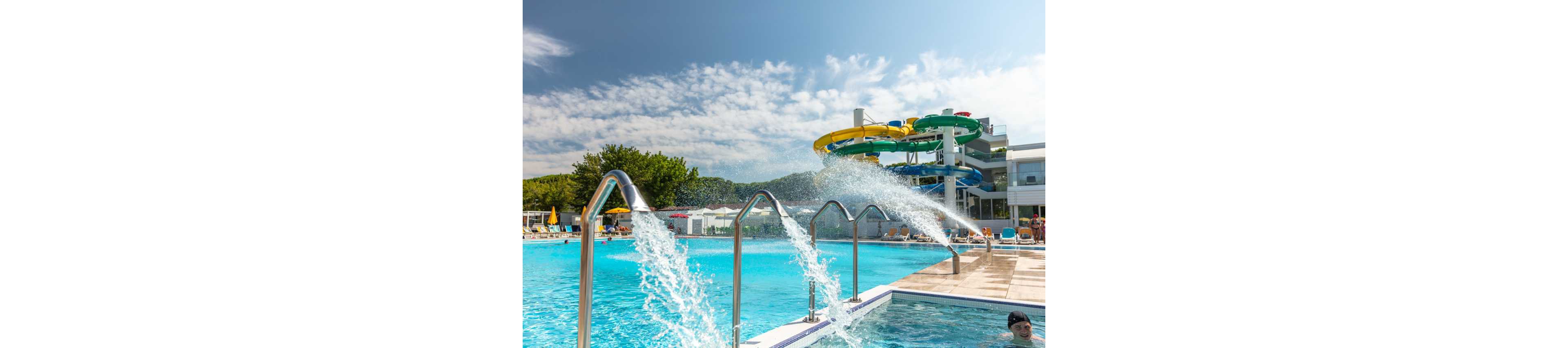A large outdoor swimming pool features several metal fountains spraying arcs of water into the bright blue pool. In the background, colorful twisting water slides rise above the pool complex under a partly cloudy sky. Sun loungers and umbrellas line the pool deck, suggesting a resort or water park setting. A few swimmers enjoy the water, creating a relaxed and playful summer atmosphere.