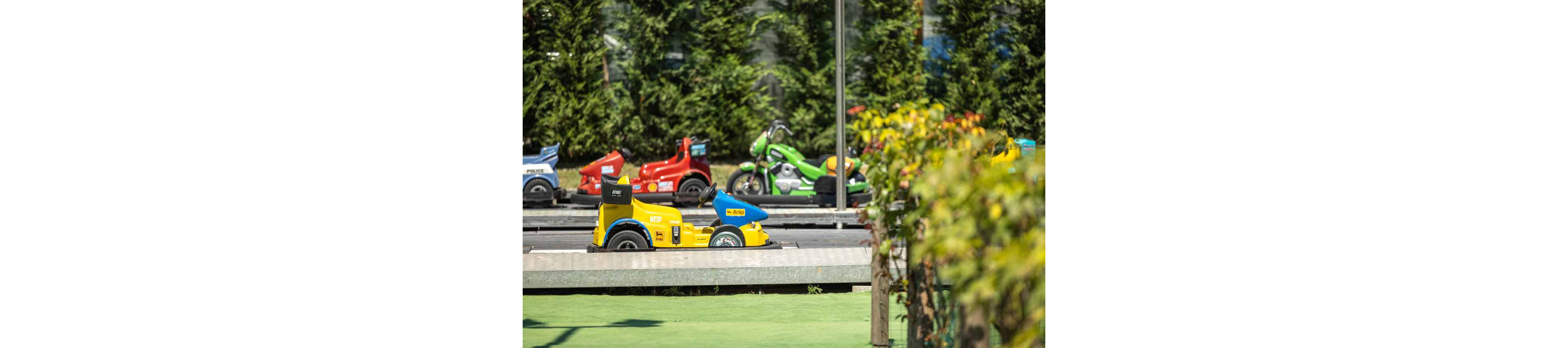 A bright yellow and blue toy vehicle is parked on a small outdoor track. Behind it, several other colorful toy cars and motorcycles are visible in a row. The scene is set in a sunny play area with green trees and plants in the background. The overall atmosphere feels playful and family-friendly.