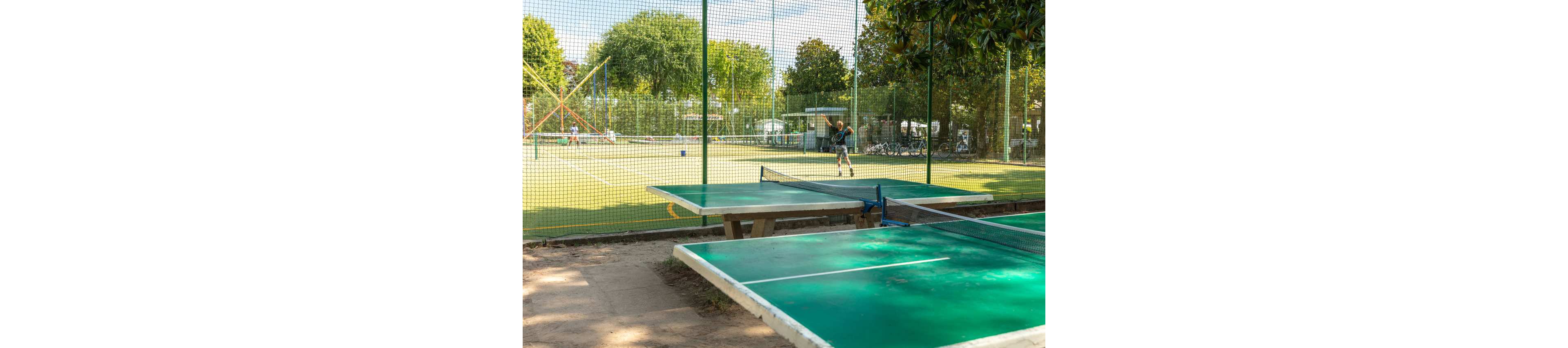 The image shows several green outdoor ping pong tables in the foreground. Behind them, a fenced tennis court is visible with a player in action. The scene is set in a sunny recreational park with lush green trees and sports facilities. The overall atmosphere is relaxed and active, suggesting a community sports area.