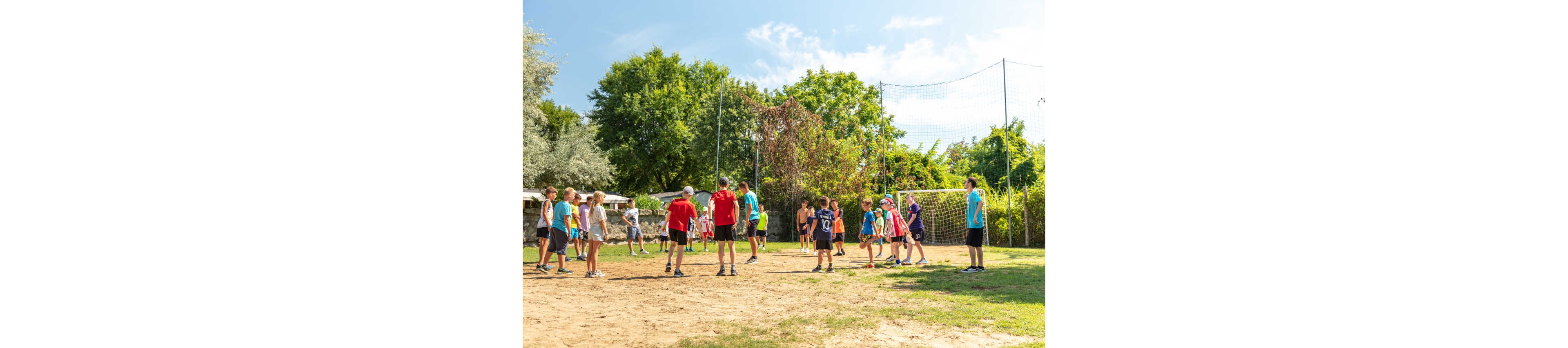 A group of children and young teens gather on an outdoor sand court to play volleyball. They stand on either side of a net, preparing for the next play under a bright blue sky. Lush green trees and grass surround the court, giving the scene a relaxed summer camp atmosphere. The kids wear colorful casual sportswear, suggesting an active and fun recreational activity.