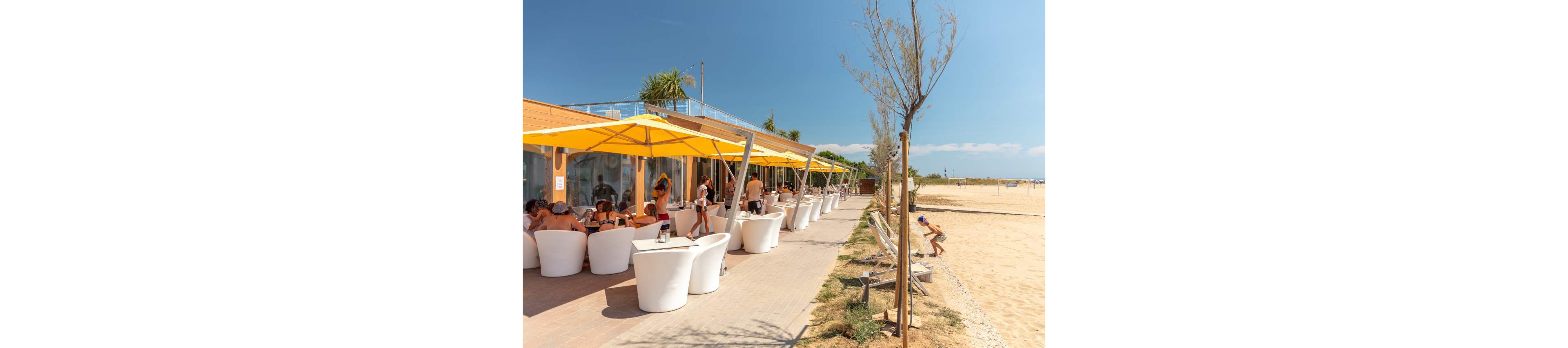 An open-air beachfront café features rows of white chairs and tables shaded by bright yellow umbrellas. People sit and socialize along the terrace while others walk nearby in swimwear. The café faces a wide sandy beach under a clear blue sky. Sparse trees and beach access paths frame the relaxed seaside setting.