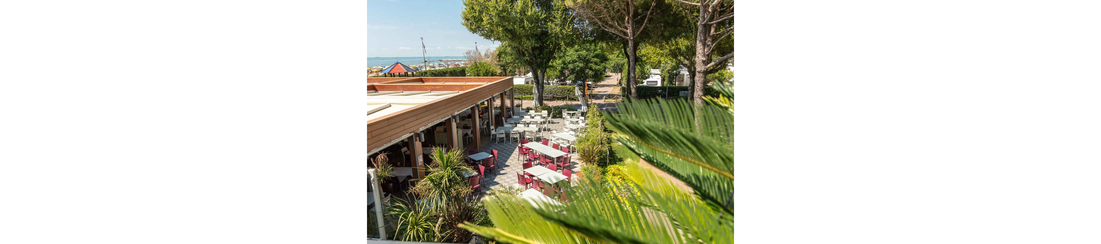 An open-air restaurant terrace is set with empty tables and chairs surrounded by lush greenery. The dining area is partially covered by a wooden structure and opens onto a paved patio. Tall trees and tropical plants frame the scene, with a glimpse of a sandy beach and calm sea in the background. The bright daylight and clear sky create a relaxed, vacation atmosphere.