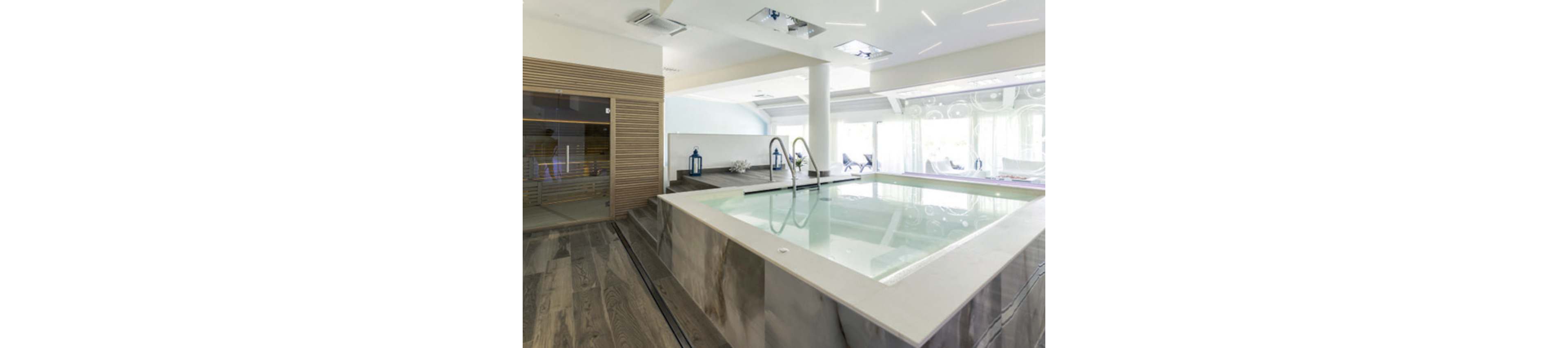 A modern indoor spa pool is featured in a bright, contemporary wellness space. The rectangular pool is surrounded by sleek stone and wood finishes, with metal handrails leading into the water. Large windows with sheer curtains allow natural light to fill the room and reveal lounge chairs in the background. A wooden sauna room and decorative lanterns enhance the relaxing, luxurious atmosphere.