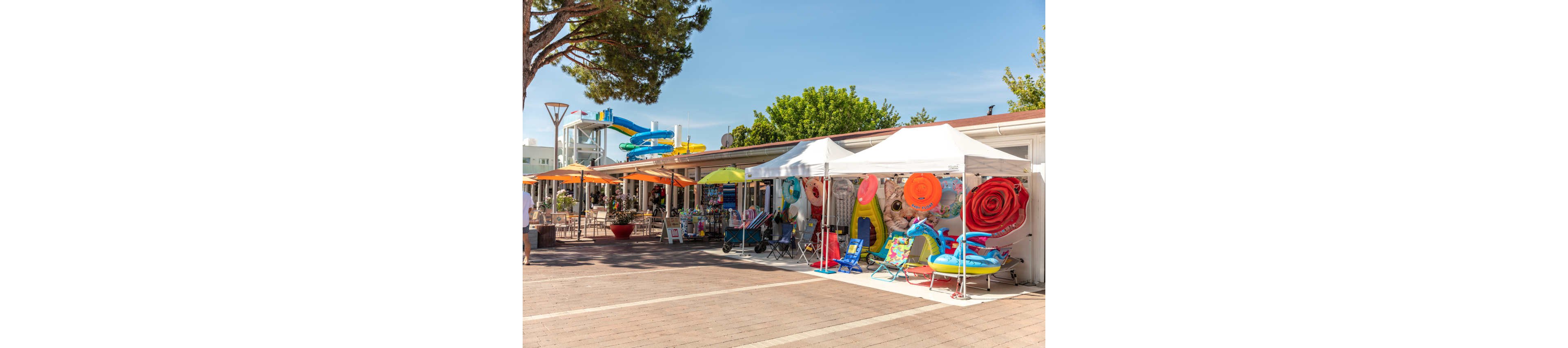 Colorful outdoor stalls display inflatable pool toys, beach chairs, and summer accessories. White canopies line a paved walkway beside a low building under a clear blue sky. In the background, bright water slides and umbrellas suggest a nearby water park or resort area. The scene feels sunny, relaxed, and geared toward vacationers and families.