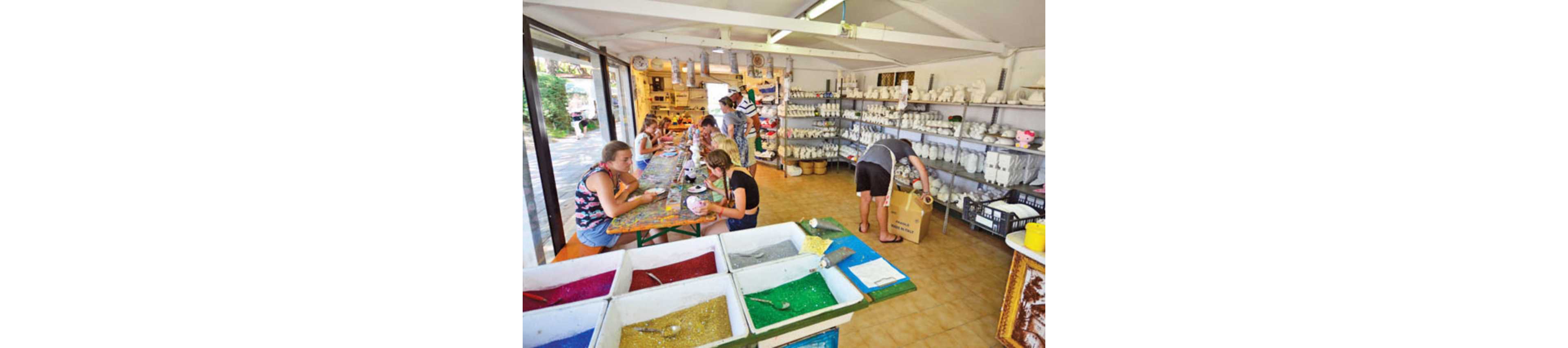 Several people sit at a long table painting ceramic objects inside a bright craft studio. Shelves along the walls are filled with unpainted pottery and figurines. In the foreground, open bins of colorful sand or glitter add vibrant texture to the scene. One person stands near the shelves handling a box while others focus on their creative projects.