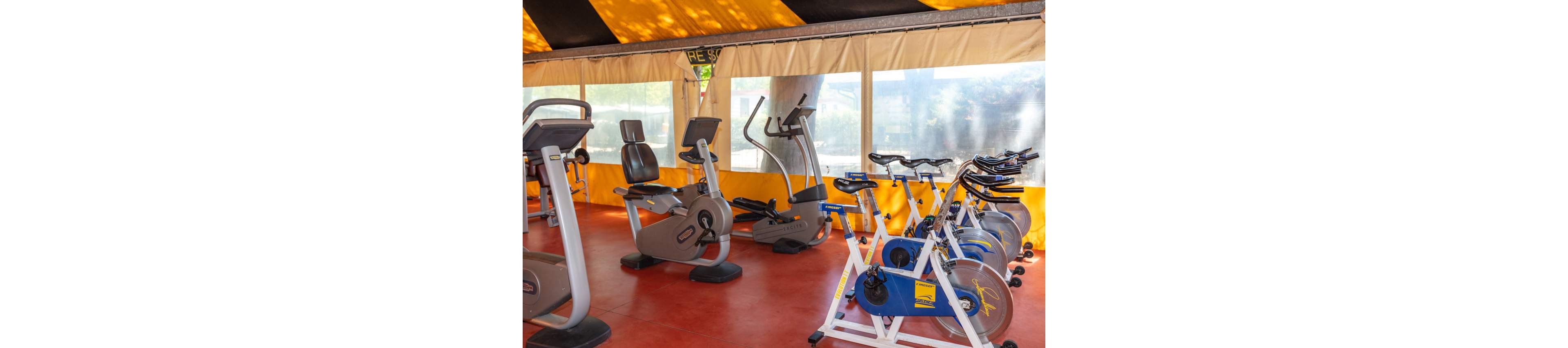 A small indoor gym features several exercise bikes and cardio machines arranged on a red floor. Stationary bikes and recumbent bikes are positioned near large windows that let in natural light. The space is covered by a yellow and black tent-like ceiling with beige curtains along the sides. The gym appears clean, bright, and ready for individual workouts.