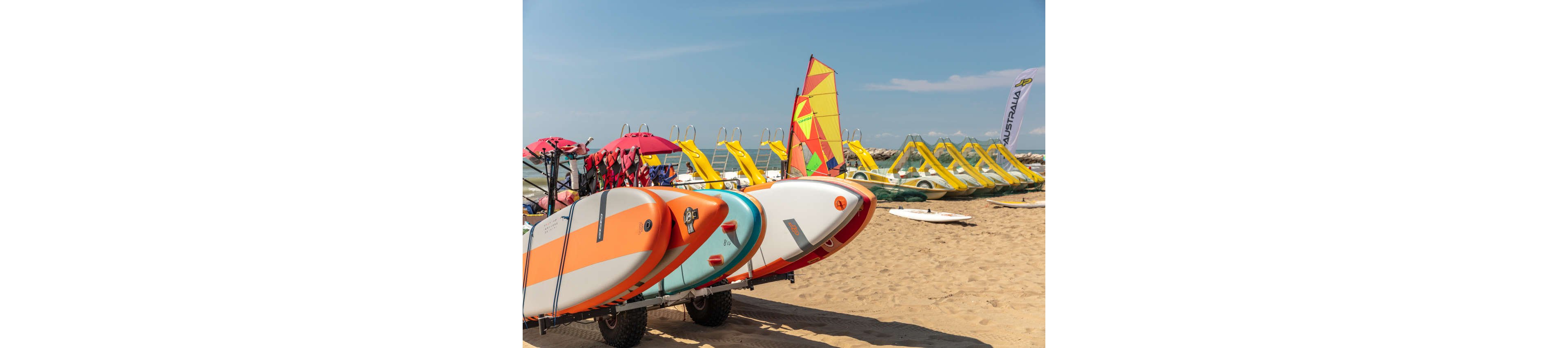 Several stand-up paddleboards are stacked on a trolley in the foreground of a sandy beach. Behind them, a row of bright yellow pedal boats lines the shoreline next to a colorful sailboard. The sea is calm under a clear blue sky, suggesting a warm summer day. A beach flag and umbrellas indicate a recreational rental area for water sports.