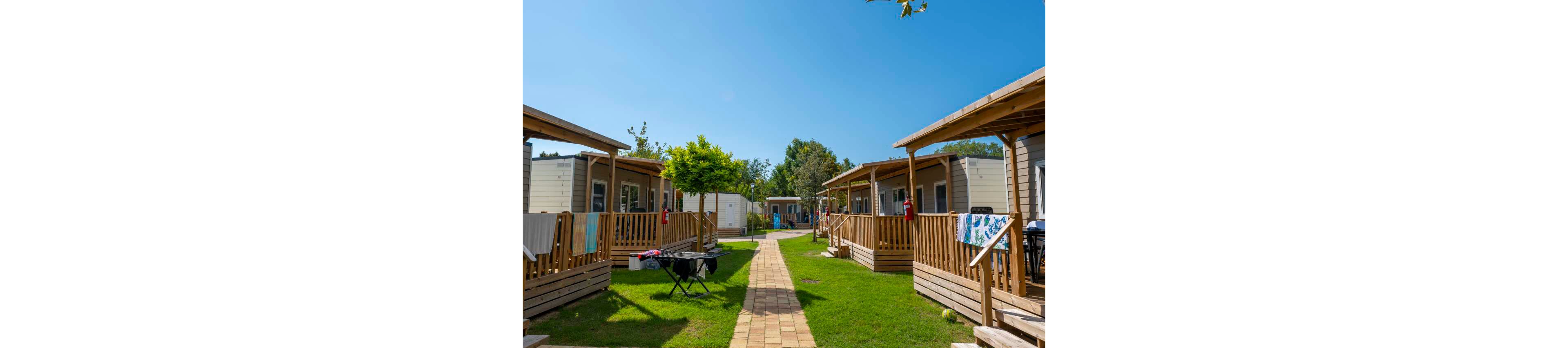 A row of modern mobile cabins with wooden porches lines a central walkway in a holiday park. Bright green grass and a clear blue sky create a warm, summery atmosphere. Towels and clothes hang on railings, and outdoor furniture is set up on the porches, suggesting vacation relaxation. Trees and additional cabins are visible in the background, enhancing the sense of a peaceful resort community.
