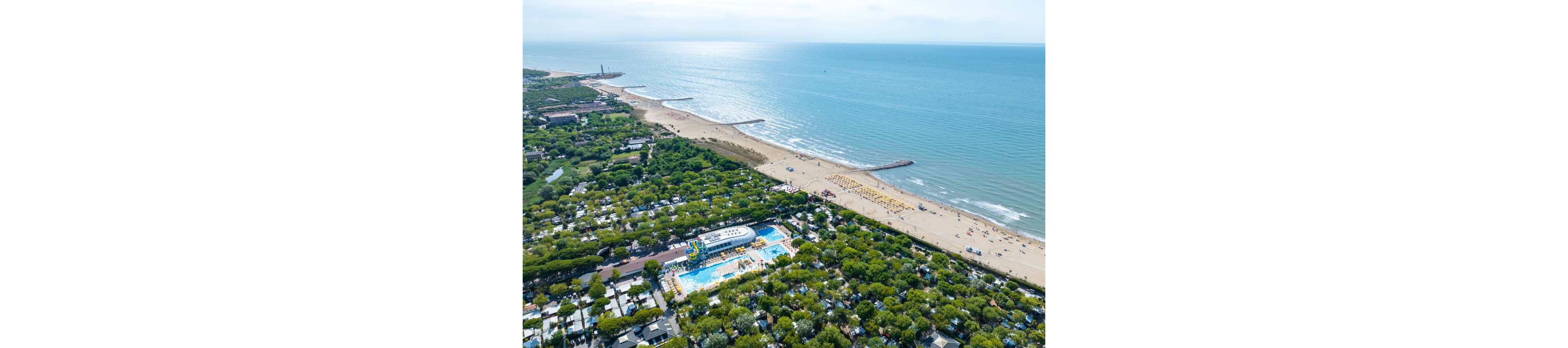 A wide aerial view shows a coastal resort town bordered by a sandy beach and calm sea. In the foreground, a large swimming pool complex with water slides is surrounded by dense green trees and numerous small buildings. The shoreline stretches into the distance with organized rows of beach umbrellas and sun loungers. The overall scene suggests a popular holiday destination with recreational facilities and natural seaside scenery.