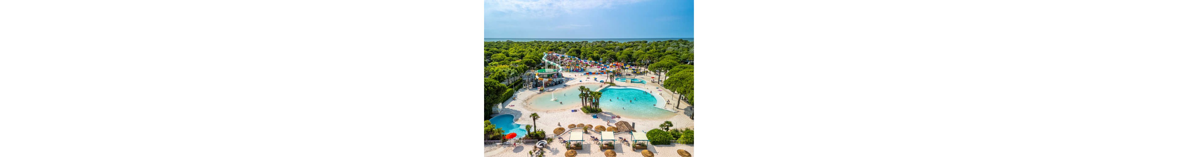 A large tropical-themed water park is seen from above, featuring expansive lagoon-style pools with palm trees and sandy-style edges. Colorful water slides and play structures dominate the center, surrounded by lush green trees and vegetation. People relax on loungers under shaded cabanas and swim in the shallow turquoise water. The scene conveys a sunny, family-friendly vacation atmosphere near the coastline.