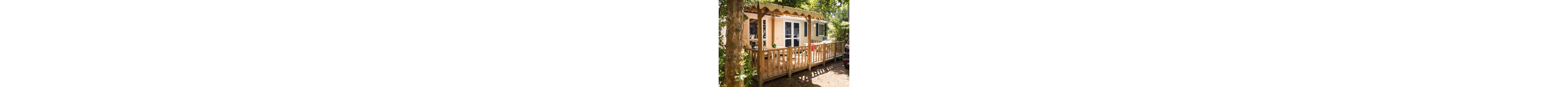 A light-colored mobile home with a wooden terrace is set among tall trees in a sunny outdoor campsite. The terrace features a railing and a partial awning providing shade near the entrance. Sunlight filters through the leaves, casting dappled shadows on the deck and ground. The scene suggests a peaceful vacation setting in nature.