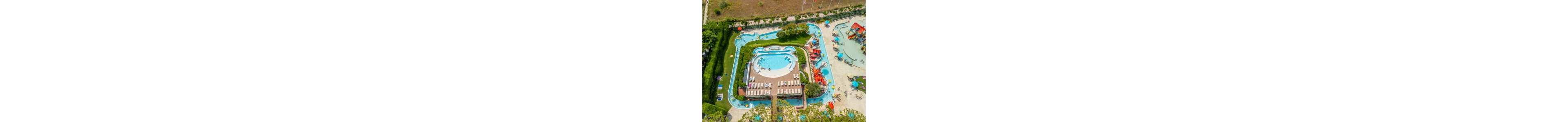A large resort-style water park is captured from above, showing a central pool surrounded by lounge chairs and umbrellas. A lazy river winds around the main pool area, with people relaxing in the water and on sunbeds. Colorful water slides and play structures are visible to the side, adding a sense of fun and activity. Lush greenery and pathways frame the aquatic attractions, creating a vibrant vacation atmosphere.