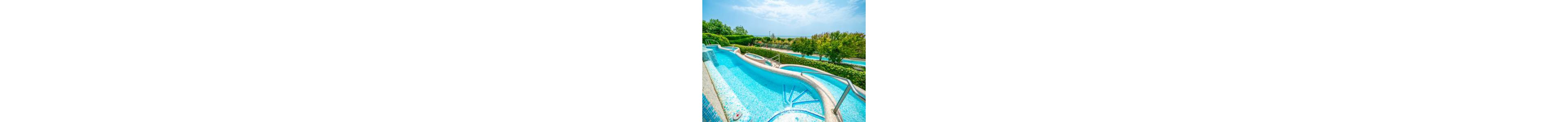 A bright blue water slide curves gently into a small pool with built-in steps. Stainless steel handrails line the slide and staircase for safety. Lush green hedges and trees surround the aquatic area, with a distant view of more water features and open landscape. The scene is captured on a sunny day under a lightly clouded sky, creating a vibrant and inviting atmosphere.