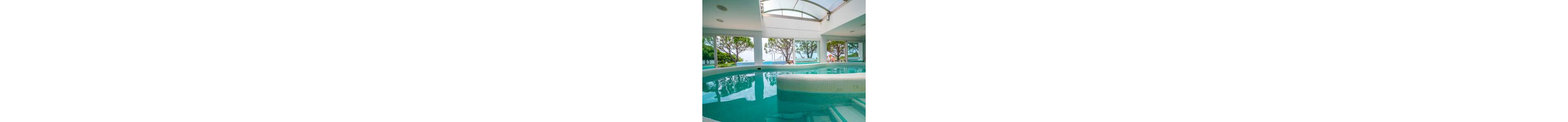 An indoor spa pool with turquoise water curves around white tiled steps in a bright room. Large windows reveal green trees and a glimpse of the sea and beach outside. Natural light fills the space, creating reflections on the calm water. The scene suggests a relaxing wellness or resort environment by the coast.
