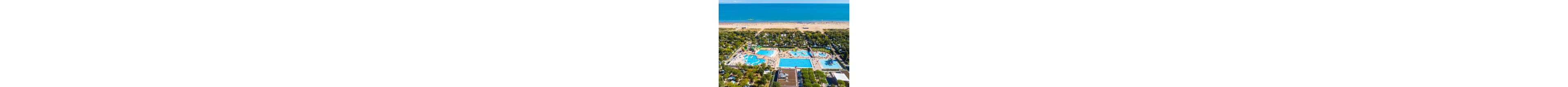 A large coastal resort with multiple outdoor swimming pools is seen from above near a sandy beach and calm blue sea. The complex includes several pools of different shapes, waterslides, and sun loungers arranged around the water areas. Green trees and vegetation surround the pools, separating them from nearby camping or accommodation areas. The bright blue water and clear sky create a vibrant, summery holiday atmosphere.