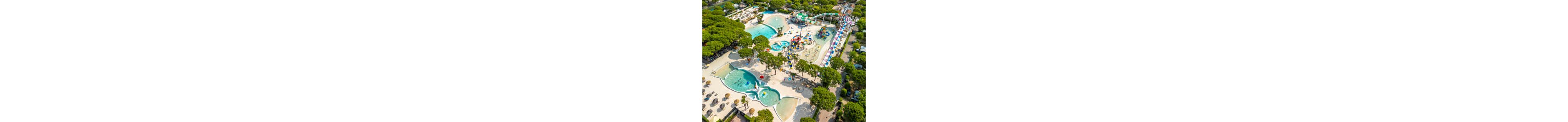 A large outdoor water park is captured from above, showing multiple pools surrounded by lush green trees. Curving water slides and colorful play structures dominate the central area, with people enjoying the attractions and sunbathing nearby. The scene includes shallow splash zones, deeper swimming areas, and winding slides that add dynamic movement. Sun loungers and umbrellas line the pool edges, creating a relaxed holiday atmosphere in a resort-like setting.