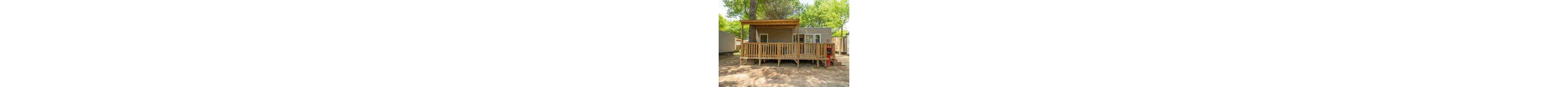 A small wooden cabin with a spacious front deck sits beneath tall green trees. The structure features a covered porch area with railing and an open section for outdoor seating. Large windows and a glass door allow natural light into the interior. The cabin is surrounded by dirt ground and neighboring mobile homes, suggesting a campground or holiday park setting.