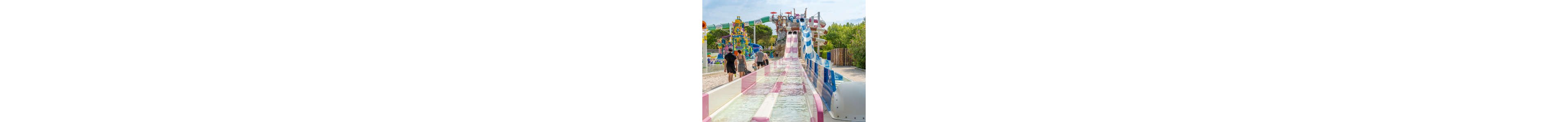 A set of colorful water slides stretches toward a splash pool in a busy outdoor water park. Several people in swimwear walk along the side of the attraction, heading toward the rides. In the background, additional play structures with sprays and buckets add to the lively scene. Lush green trees and bright daylight create a cheerful, summery atmosphere.