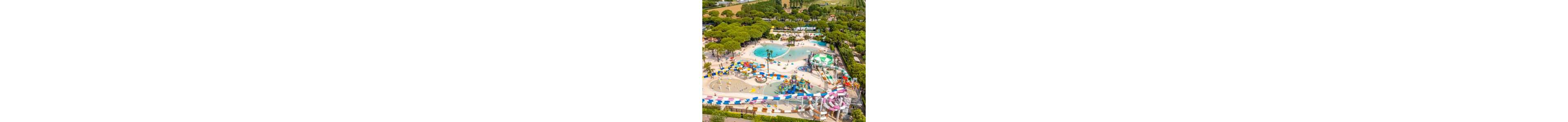 A large outdoor water park is captured from above, showing multiple pools and winding water slides. The complex features splash zones, shallow play areas, and a central water playground with fountains and sprayers. Brightly colored slides, umbrellas, and play structures stand out against the surrounding green trees and landscaped grounds. The scene suggests a family-friendly recreational area on a sunny day.
