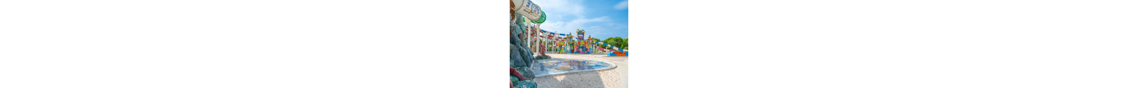A vibrant outdoor water park features multiple colorful slides and play structures under a bright blue sky. The foreground shows a shallow splash pad area with playful designs and sandy ground. In the background, tall twisting water slides and towers are surrounded by lush green trees. The scene appears calm with no people visible, emphasizing the fun, family-friendly environment.