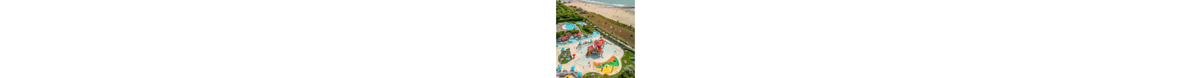 A vibrant waterpark with colorful slides and splash structures is captured from above near a sandy beach. Families and children enjoy shallow pools, sun loungers, and umbrellas around the main play area. Additional swimming pools and landscaped green spaces surround the central attraction. In the background, people relax and walk along the wide shoreline by the sea.