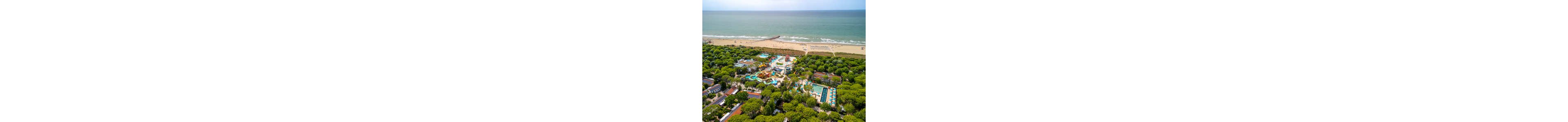 A large seaside resort with multiple swimming pools and water slides is seen from above. The complex is surrounded by dense green trees and low-rise holiday buildings. In the background, a wide sandy beach stretches along the shoreline, meeting calm ocean waves. Colorful pool areas, sun loungers, and pathways create a vibrant vacation atmosphere in a natural coastal setting.