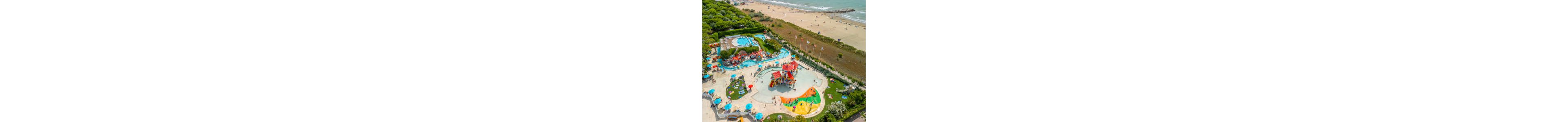 A colorful water park with pools and slides is captured from above near a sandy beach. Families and children relax on sun loungers and play in shallow splash areas surrounded by umbrellas. The resort is bordered by green trees and landscaped gardens, with walking paths connecting different pool zones. In the background, people stroll and sunbathe along the shoreline beside the calm sea.
