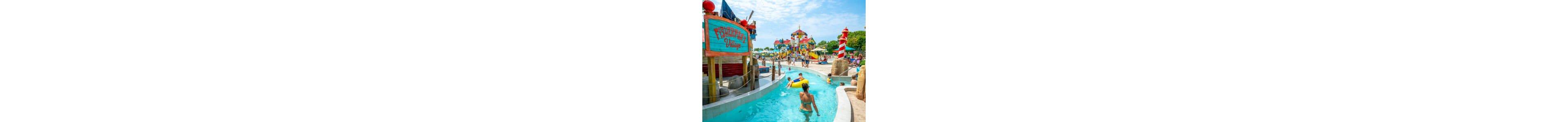 Families float along a bright blue lazy river in a colorful outdoor waterpark. A large wooden sign reading "Fisherman's Village" stands beside the water, while children and adults play in the background. The scene includes vibrant slides, towers, and a striped lighthouse structure under a sunny sky. The overall atmosphere is fun, energetic, and summery.