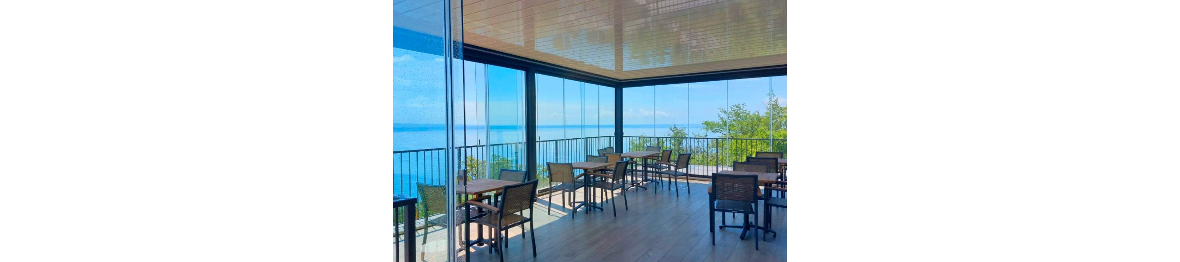 An open-air café terrace with glass walls overlooks a calm blue sea. Several empty tables and chairs are arranged on a wooden floor under a sleek covered roof. Metal railings frame the edge of the terrace, enhancing the elevated coastal view. Bright daylight and clear skies create a relaxed, inviting atmosphere for outdoor dining.