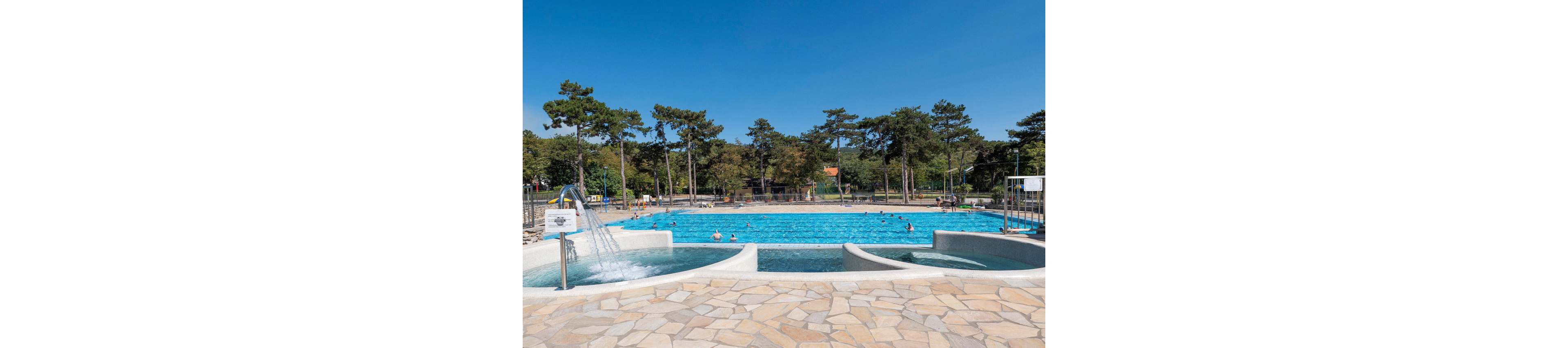 An outdoor public swimming pool is shown under a clear blue sky, surrounded by tall trees and greenery. In the foreground, a shallow pool with water features and stone paving leads toward a large rectangular lap pool. Several people are swimming and relaxing in the water, enjoying the warm weather. The scene conveys a relaxed summertime atmosphere in a recreational park setting.