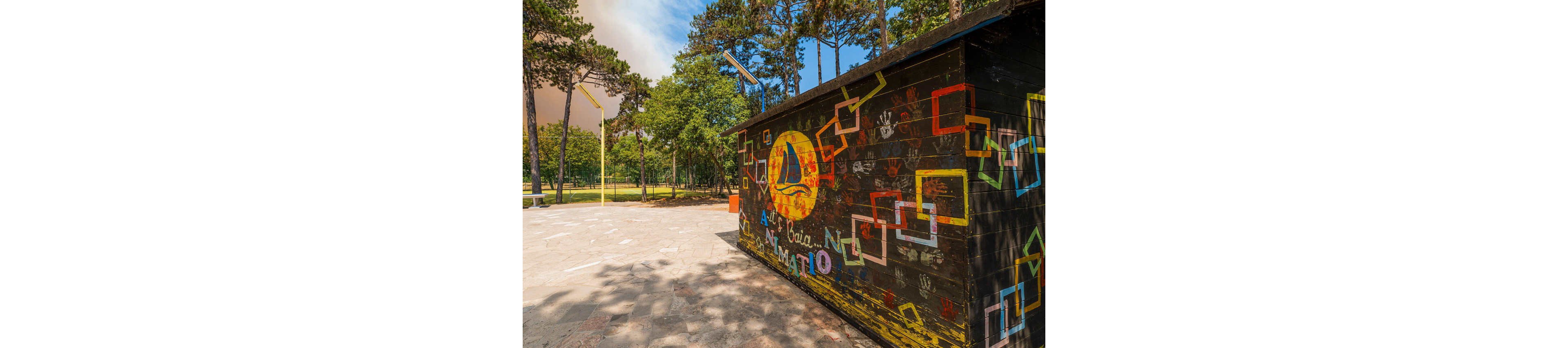 A small wooden kiosk is covered with colorful hand-painted artwork, including a large yellow circle with a sailboat and overlapping geometric shapes. The structure stands on a paved area at the edge of a green park filled with tall trees. Sunlight casts soft shadows on the ground, creating a warm and inviting outdoor atmosphere. The scene suggests a recreational or community space, possibly used for activities or information.