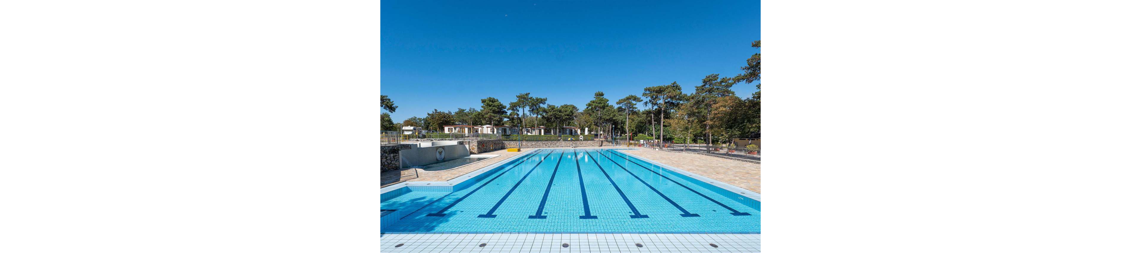 A large outdoor lap pool stretches into the distance under a bright blue sky. The pool is empty, with clear water and marked swimming lanes. Surrounding the pool are paved walkways, safety railings, and a backdrop of trees and low buildings. The scene suggests a quiet moment at a recreational or training facility in a natural setting.